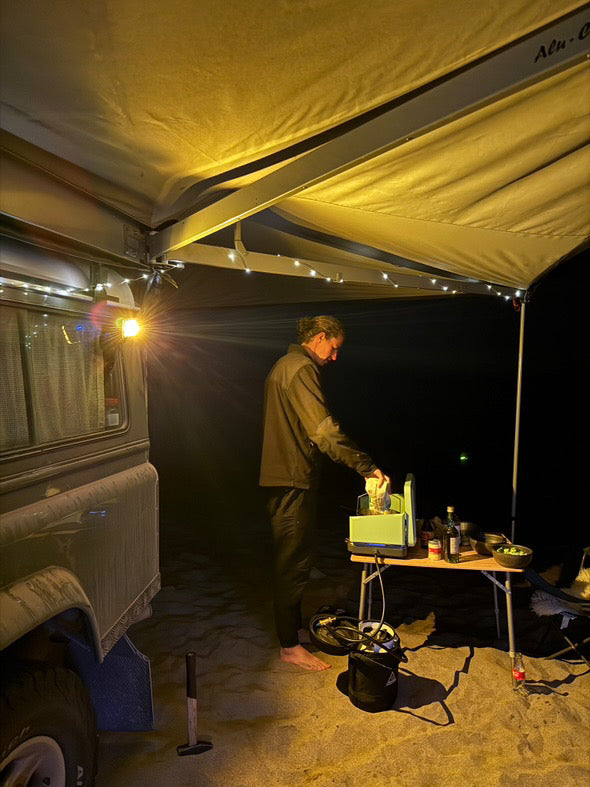 Person preparing food under a tent at night near a land rover defender with an E-Sea Light shining bright on the campsite