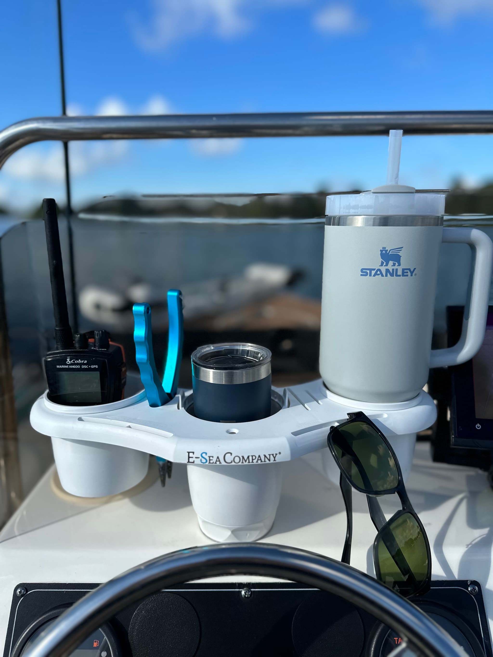 Boat dashboard with E-Sea Caddy cup holder, sunglasses, pliers, VHF radio and a Stanley mug against a blurred outdoor background.