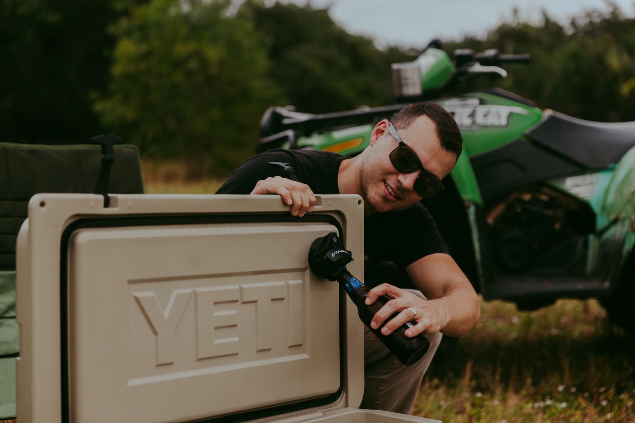 An open Yeti cooler with an E-Sea Bottle Opener suction mounted inside the lid opening a bottle at a campsite with an atv in the background